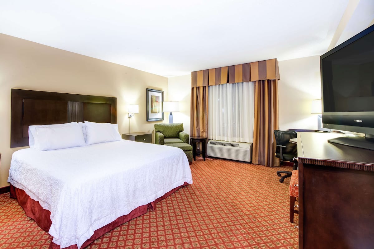 A hotel room with a large bed, a desk with a flat-screen TV, a chair, a desk lamp, and a window with curtains.