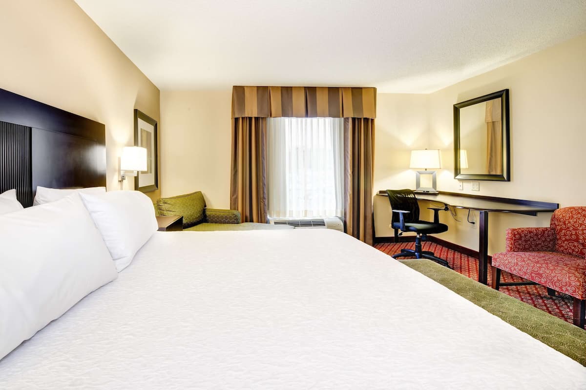 A hotel room with a large bed, a desk with a chair, a small sofa, and a window with curtains.