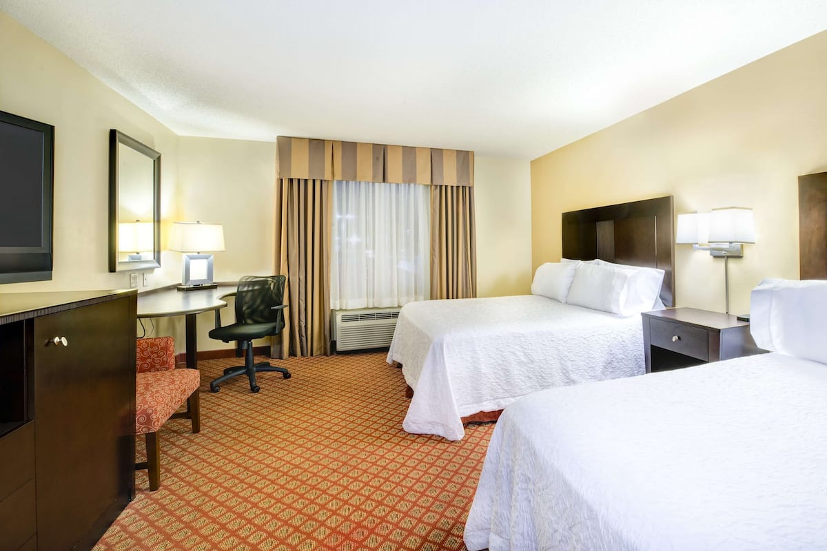 A hotel room with two beds, a desk, a chair, a TV, and a window with curtains.