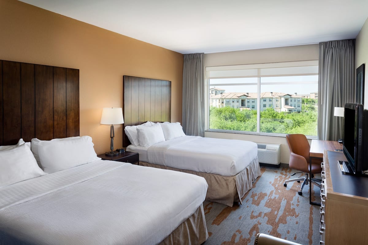 A hotel room with two beds, a desk with a chair, a television, and a large window with a view of buildings and greenery.
