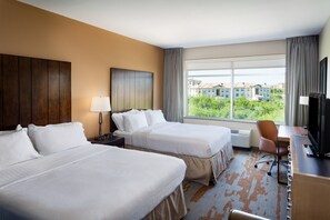 Habitación de hotel con dos camas, un escritorio con silla, televisor y ventana grande con vistas a edificios y zonas verdes.