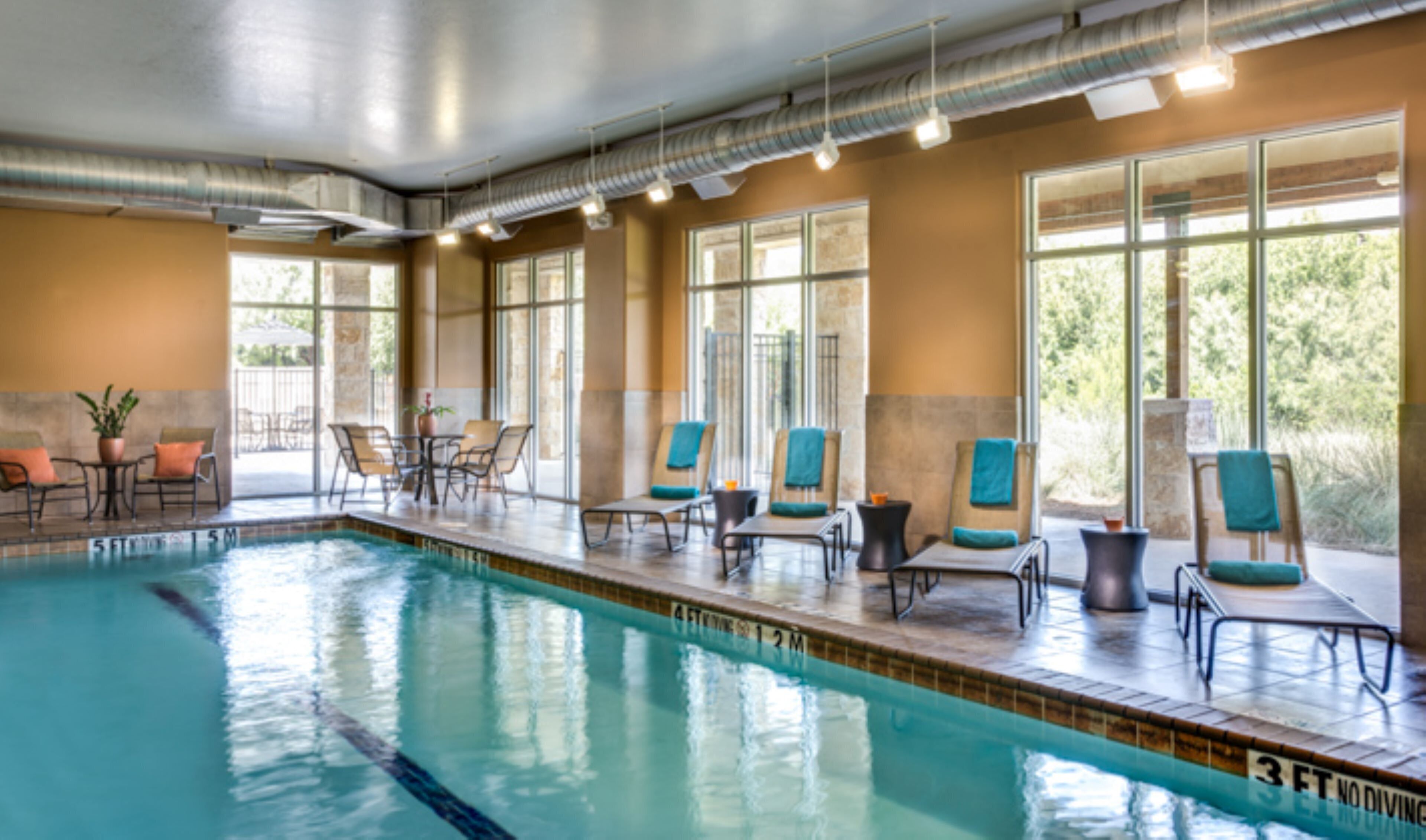 Una piscina con sillones de descanso y mesas, ventanas amplias y vistas a zonas verdes.