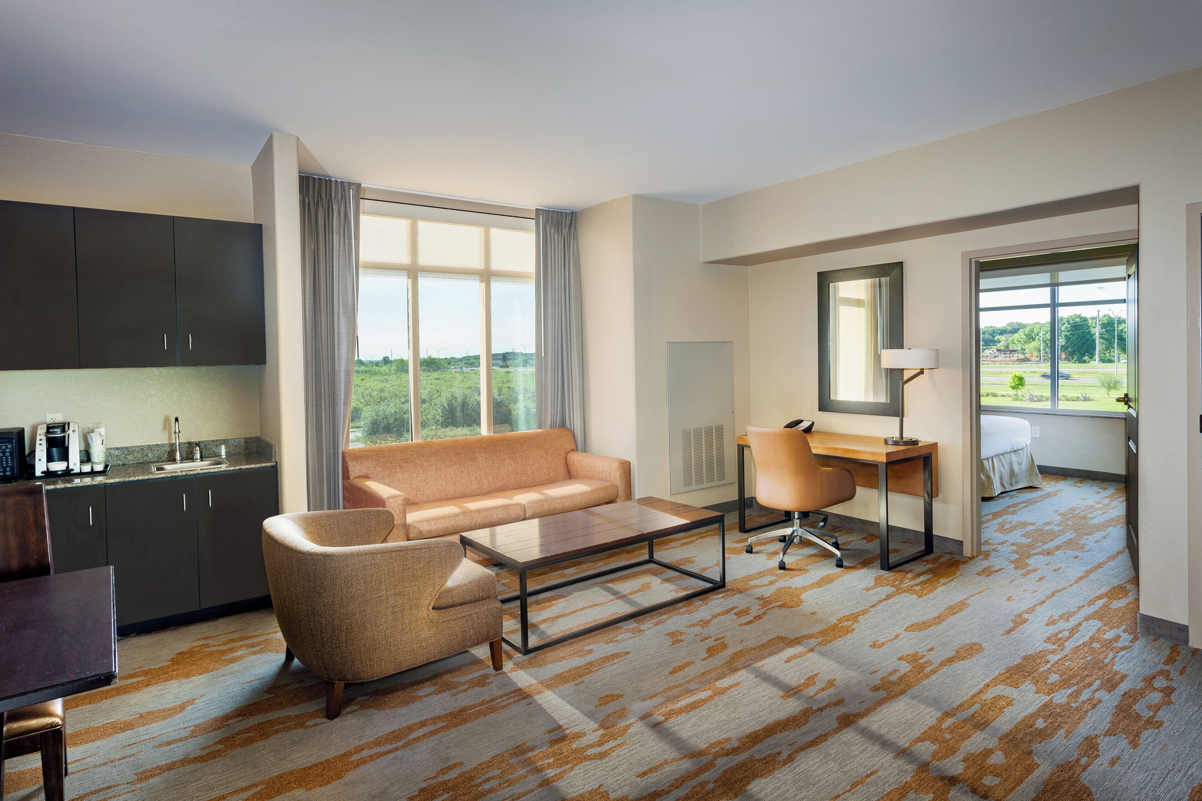 A hotel room with a sofa, a desk, a chair, a coffee machine, and a view of greenery.