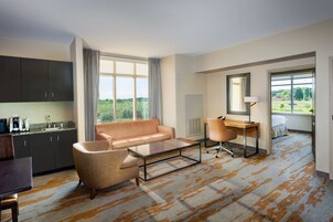 A modern hotel room with a sofa, coffee table, desk, and a view of greenery outside.