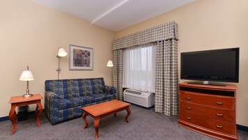 A hotel room with a sofa, a wooden coffee table, a TV on a wooden cabinet, a lamp, and a framed picture on the wall.
