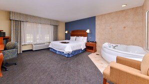 A hotel room with a large bed, a sitting area with a chair, a desk, a desk lamp, a nightstand, a bedside table, a lamp, a large window with curtains, an air conditioning unit, a carpet, and a bathtub. - Hampton Inn Evanston (Evanston)