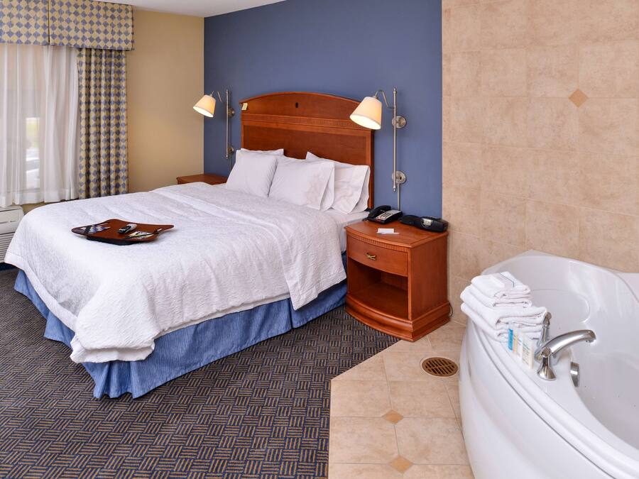 A hotel room with a large bed, a wooden nightstand, a telephone, a tray with remote controls, and a bathtub with towels.