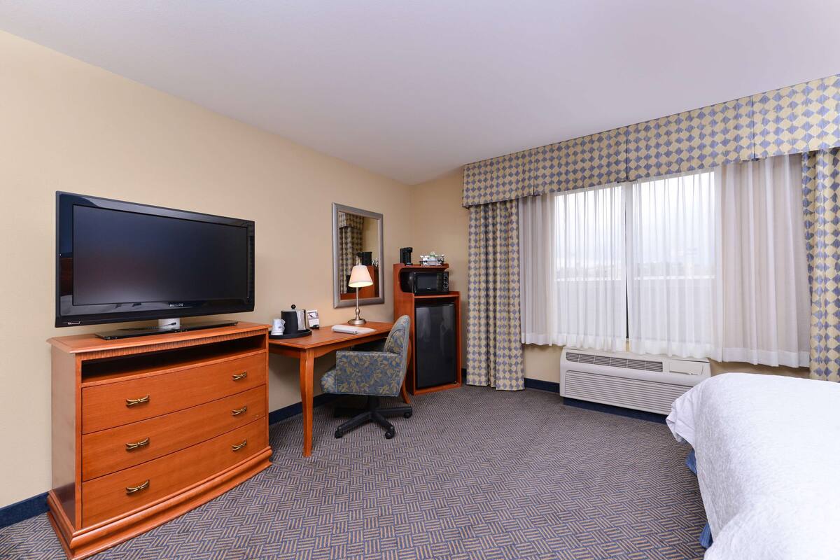 Une chambre d’hôtel équipée d’une télévision à écran plat, d’un bureau avec une chaise, d’une commode, d’un lit et d’une fenêtre avec des rideaux à motifs.