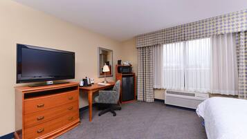 Habitación de hotel con televisión de pantalla plana, escritorio con silla, cómoda, cama y ventana con cortinas estampadas.