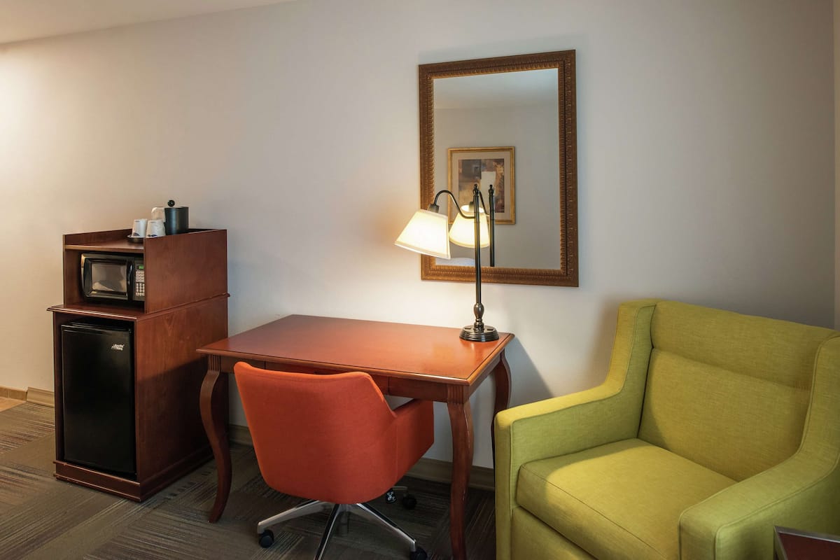 Quarto de hotel com escrivaninha de madeira, poltrona verde, micro-ondas, frigorífico e candeeiro.