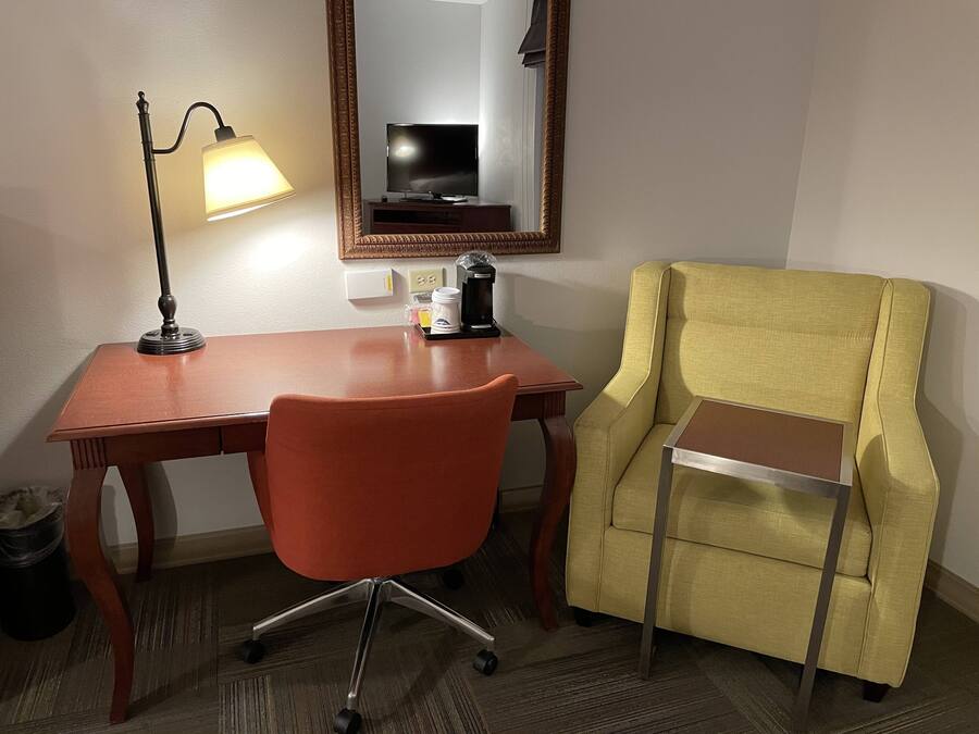 A hotel room with a wooden desk, a red office chair, a yellow armchair, a lamp, a coffee maker, and a television.