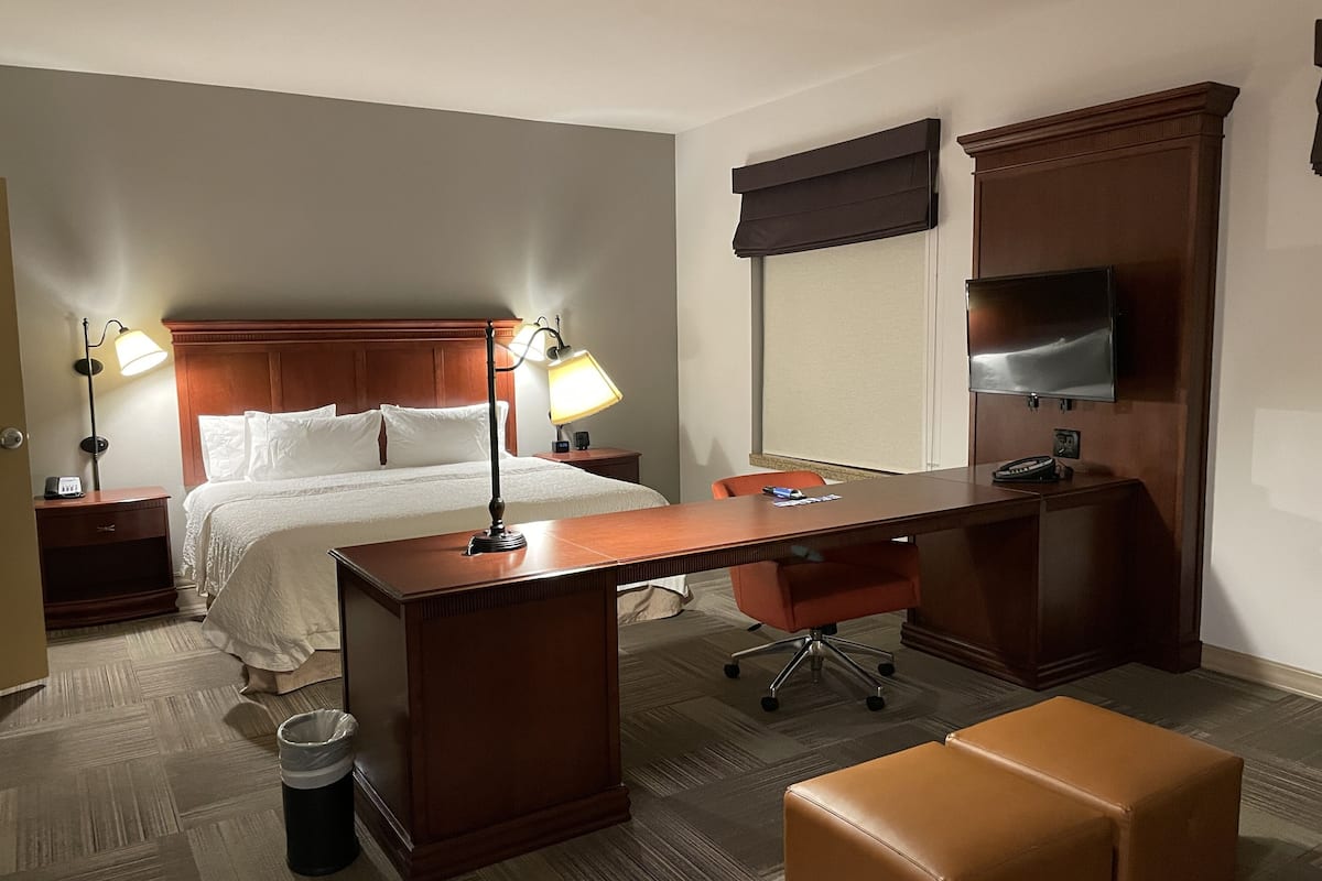 Quarto de hotel com cama, duas luminárias de cabeceira, escrivaninha com cadeira, televisão e lixeira.