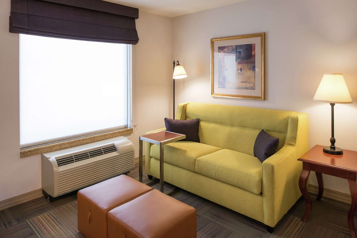 Quarto de hotel com sofá amarelo, um banquinho marrom, uma mesinha, duas luminárias de chão, um quadro na parede e uma janela com cortina escura.