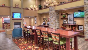 A hotel lobby with a long wooden bar table, multiple chairs with patterned cushions, a stone accent wall, and a fireplace.