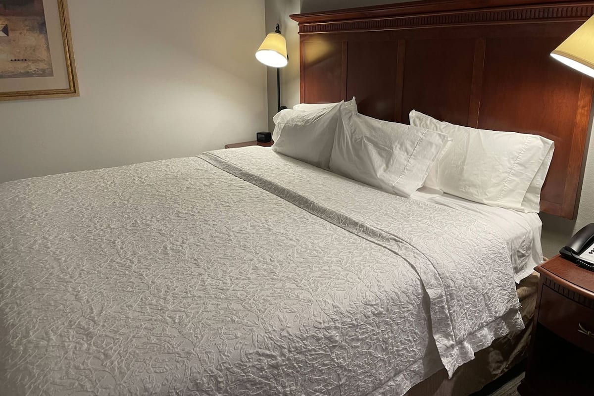 Cama bem arrumada com colcha branca texturizada, cabeceira de madeira e duas luminárias laterais.
