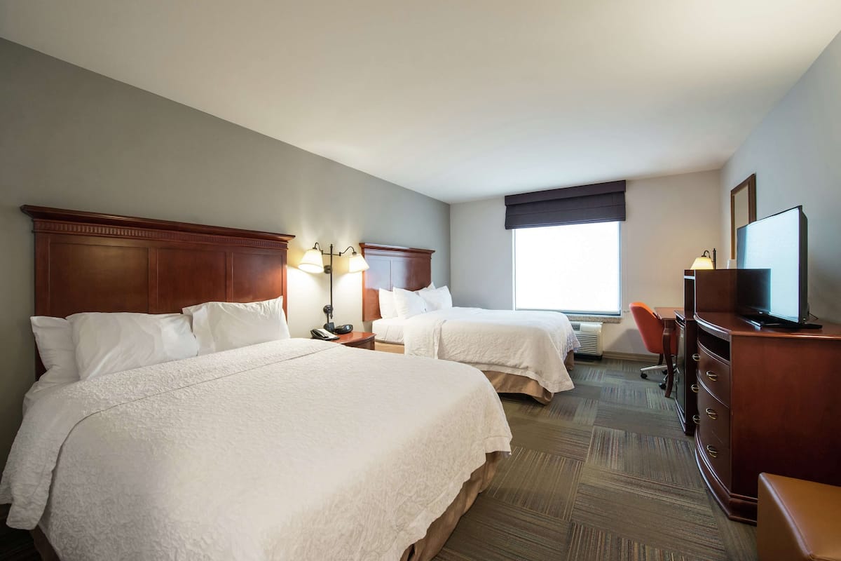 Quarto de hotel com duas camas, uma escrivaninha, uma cadeira, televisão e uma janela com vista.