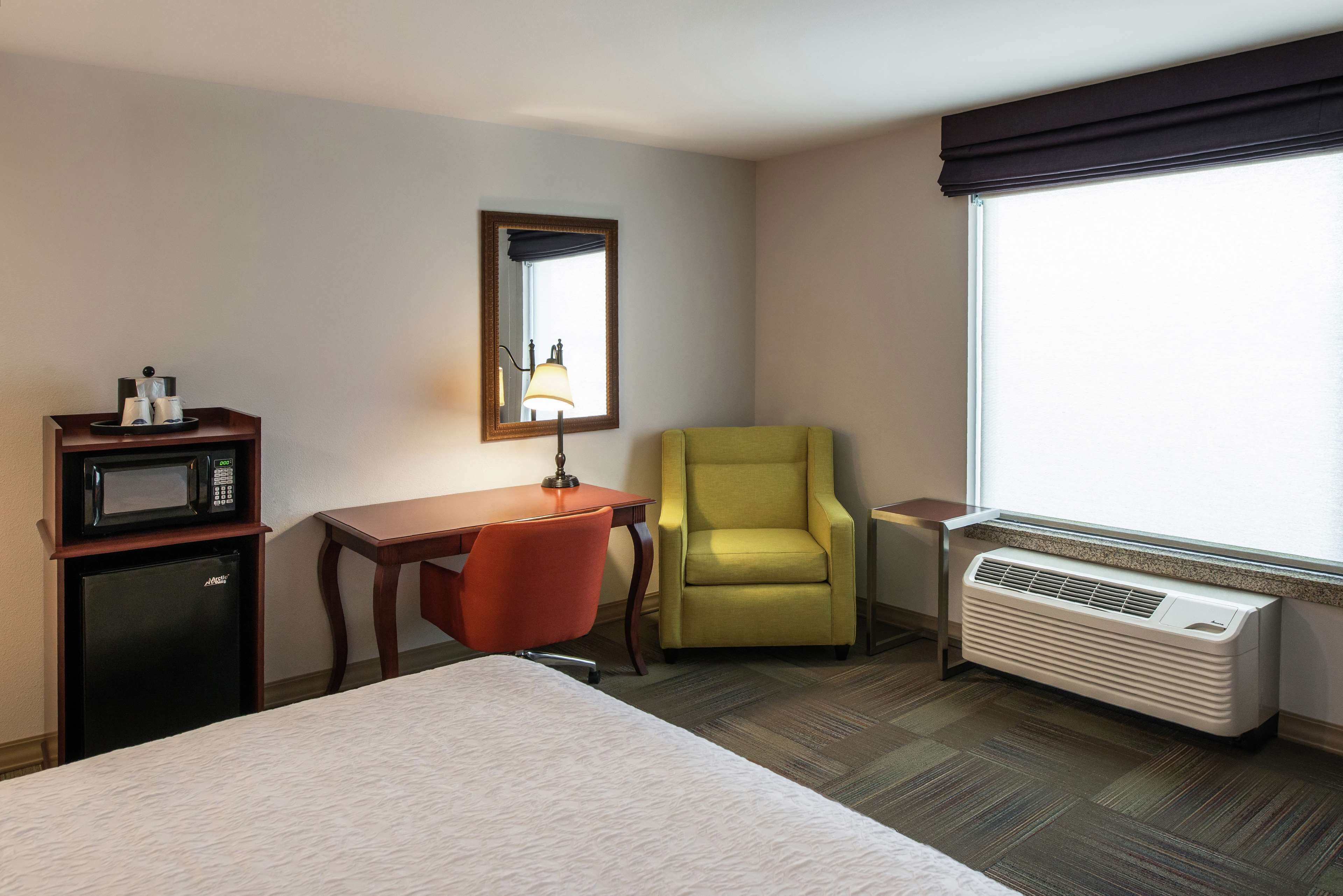 A hotel room with a bed, a desk with a microwave, a chair, a large window, and an air conditioner.