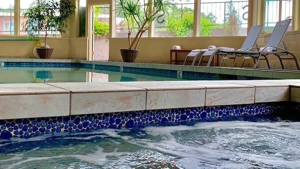 Una piscina interior con agua cristalina, bordes de azulejos y un borde de mosaico azul. Hay sillones de descanso y plantas en macetas cerca de las ventanas.
