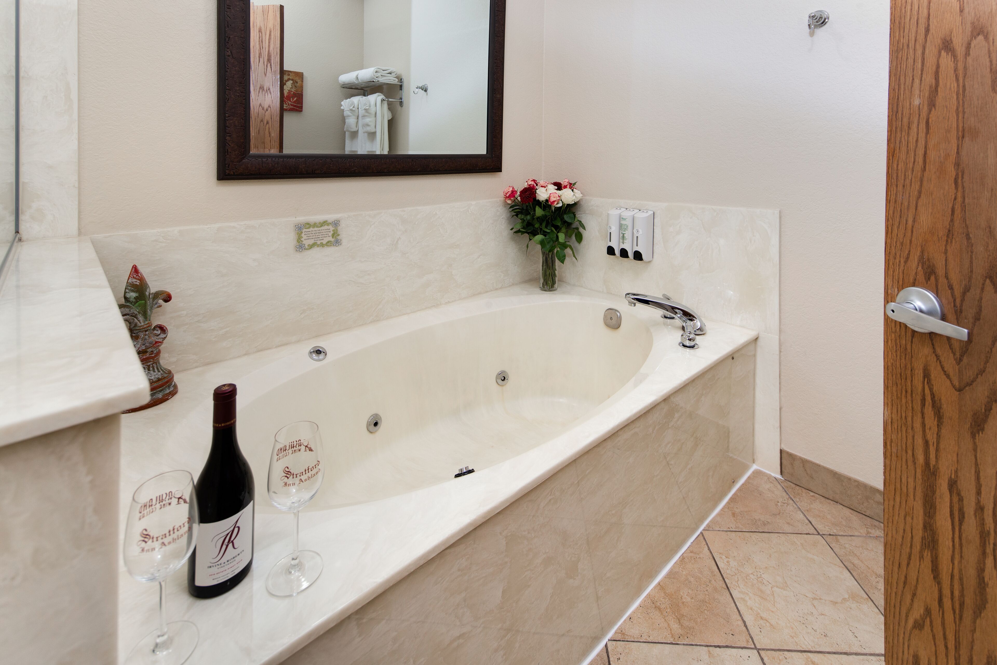 A bathroom with a large bathtub, a mirror, a vase with flowers, and a bottle of wine.
