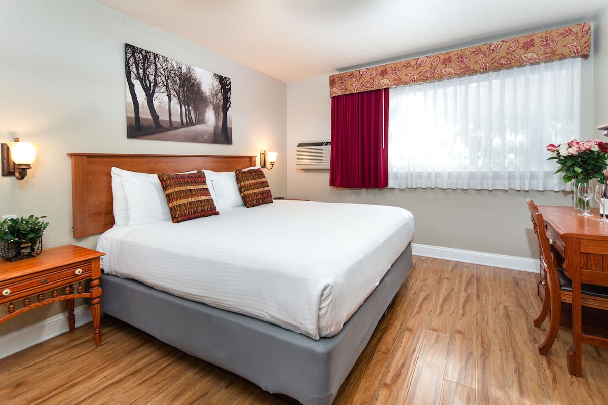 Quarto de hotel bem arrumado com uma cama grande, uma mesa de cabeceira de madeira, uma escrivaninha com cadeira e uma janela com cortinas.