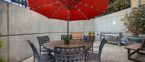 An outdoor patio with a round table and six chairs, a red umbrella, and a barbecue grill.