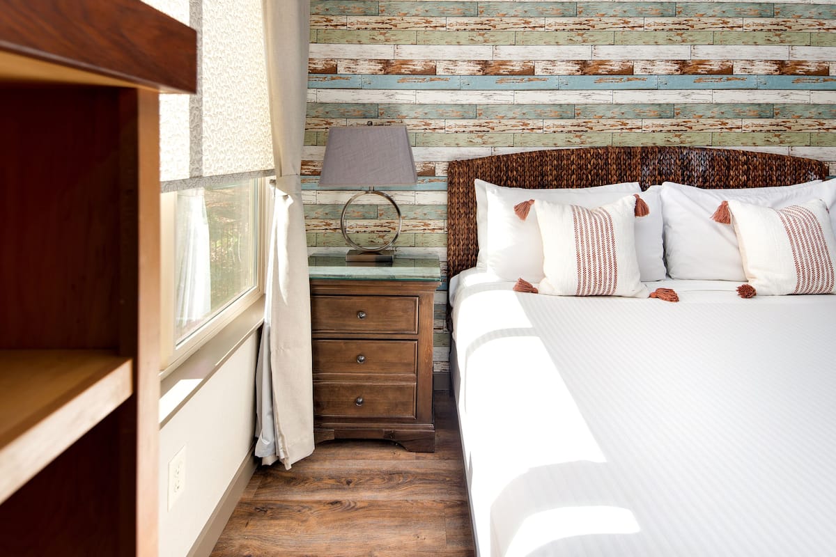 Quarto com cabeceira de madeira, mesa de cabeceira com abajur e janela com cortinas.