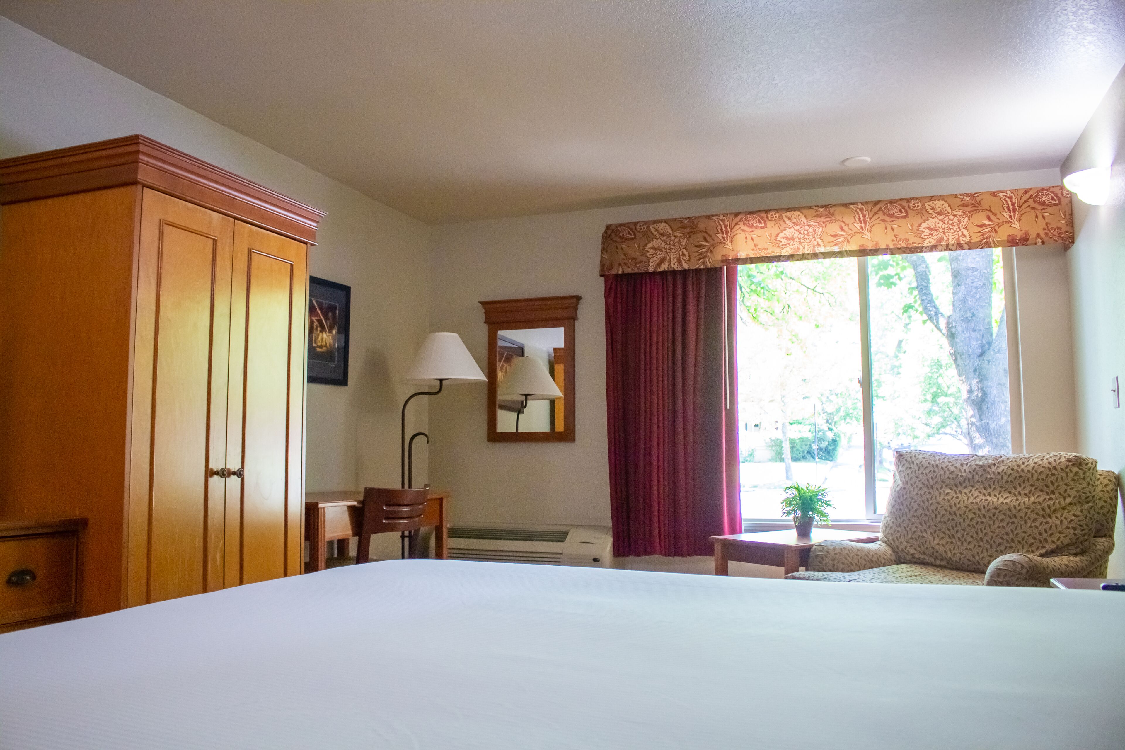 A hotel room with a large bed, a wooden wardrobe, a desk with a chair, a lamp, a window with curtains, and a sofa.