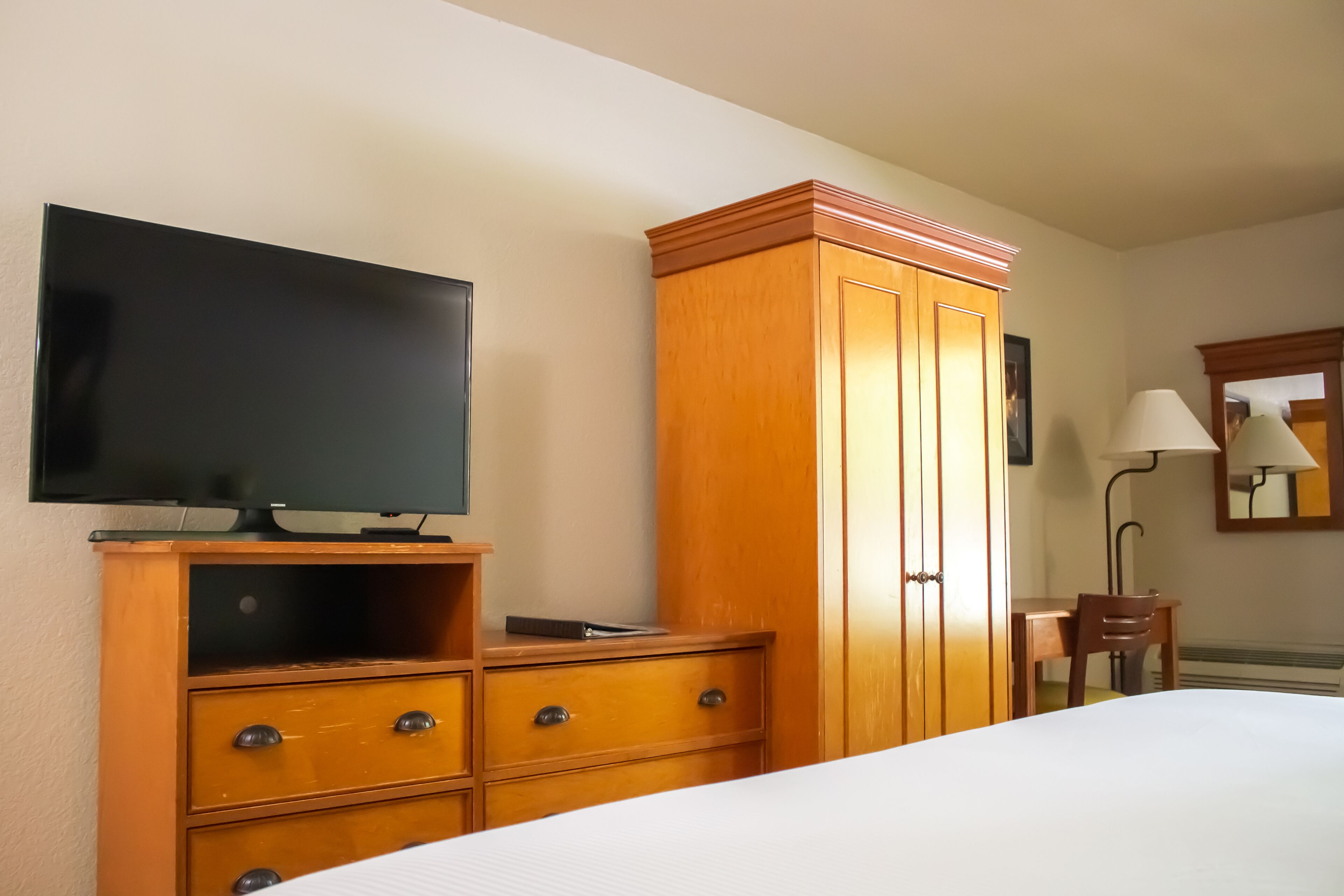 A hotel room with a bed, a wooden wardrobe, a television, a desk with a chair, and a lamp.