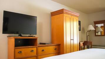 A hotel room with a bed, a wooden wardrobe, a television, a desk with a chair, and a lamp.