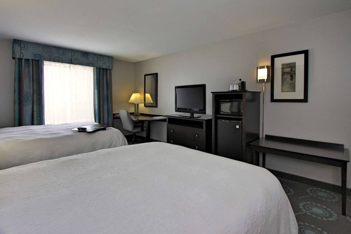 A hotel room with two beds, a desk with a computer, a chair, a lamp, a television, and a small cabinet.