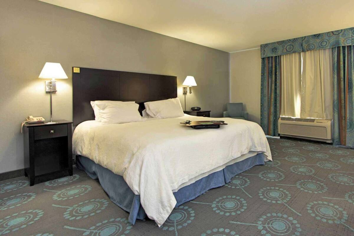 A hotel room with a bed, two bedside tables, a lamp, a chair, a window with curtains, and an air conditioning unit.