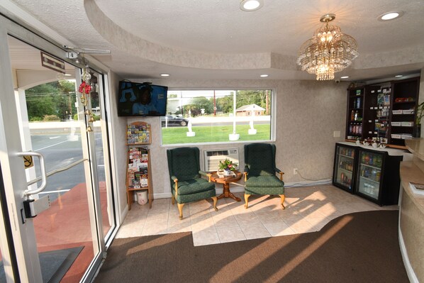 A lobby with two green armchairs, a chandelier, a refrigerator, and a television on the wall.