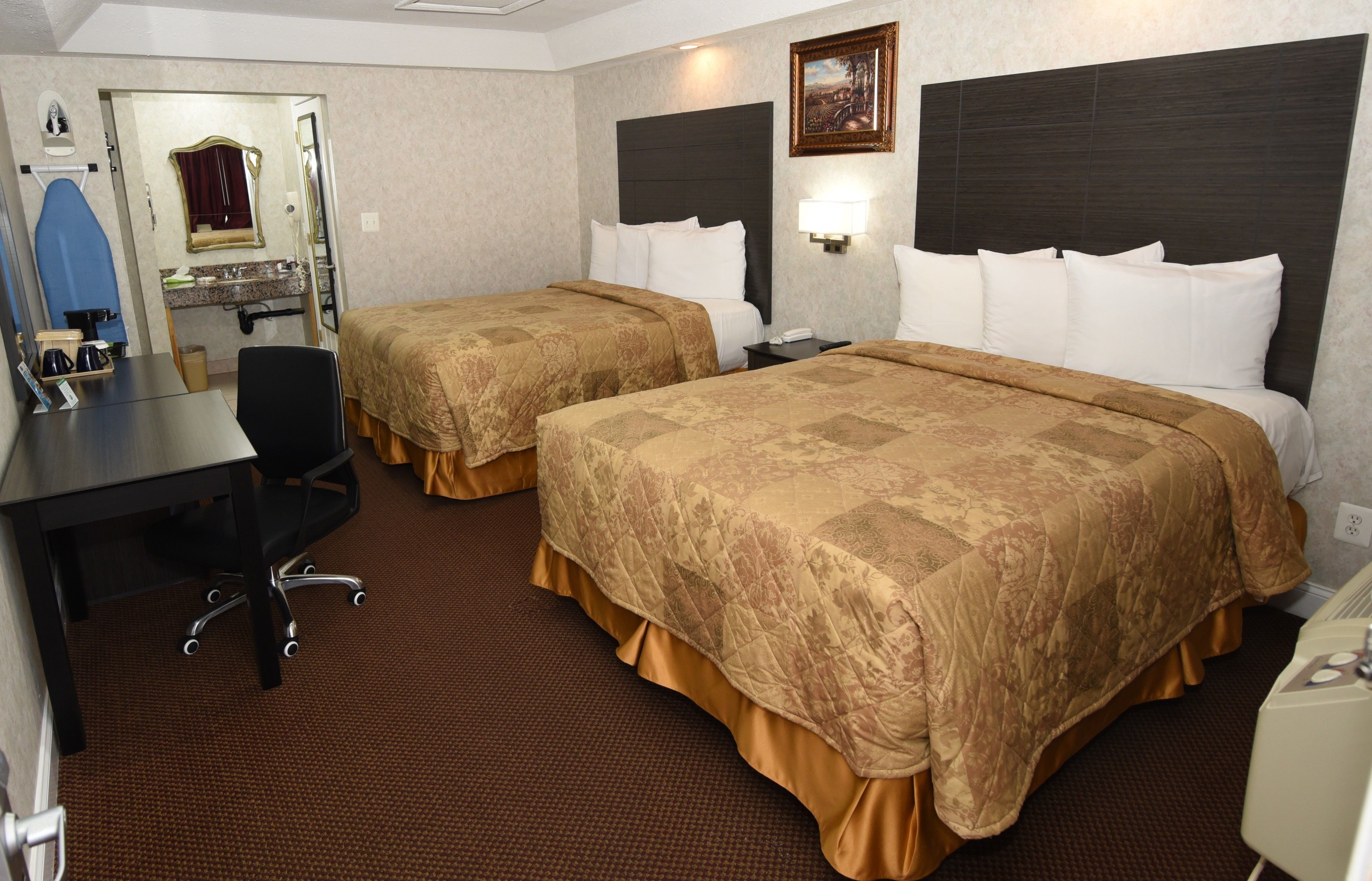 Quarto de hotel com duas camas, uma escrivaninha, uma cadeira, toucador com espelho e um quadro na parede.