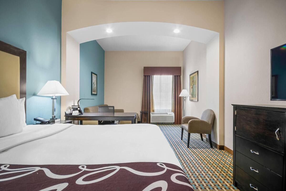A hotel room with a large bed, a desk, a chair, a dresser, and a window with curtains.