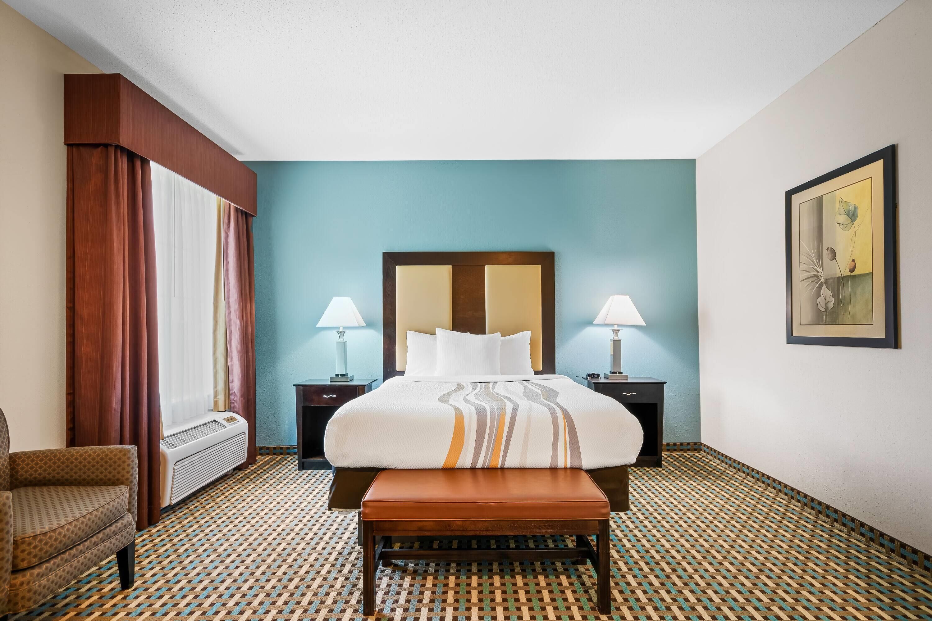 Una habitación de hotel con una cama, dos lámparas de noche, un banco, una silla y un suelo estampado.