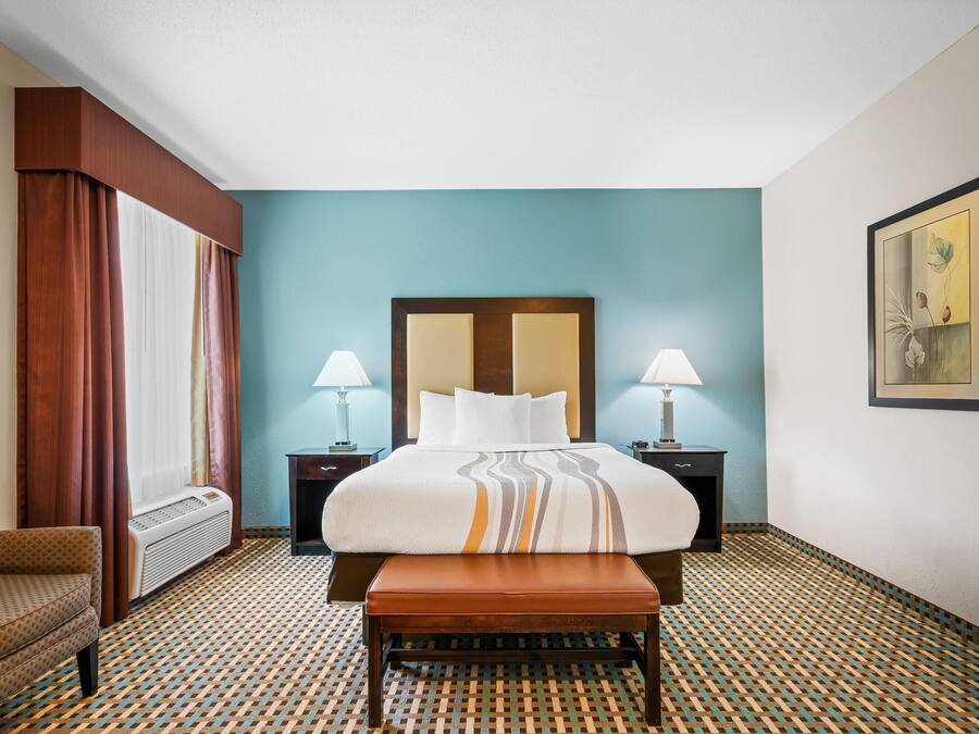 A hotel room with a bed, two bedside lamps, a bench, a chair, and a patterned floor.