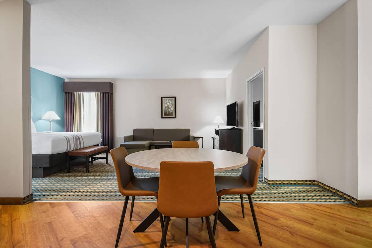 A hotel room with a round dining table, four chairs, a sofa, a bed, a TV, and a patterned rug.