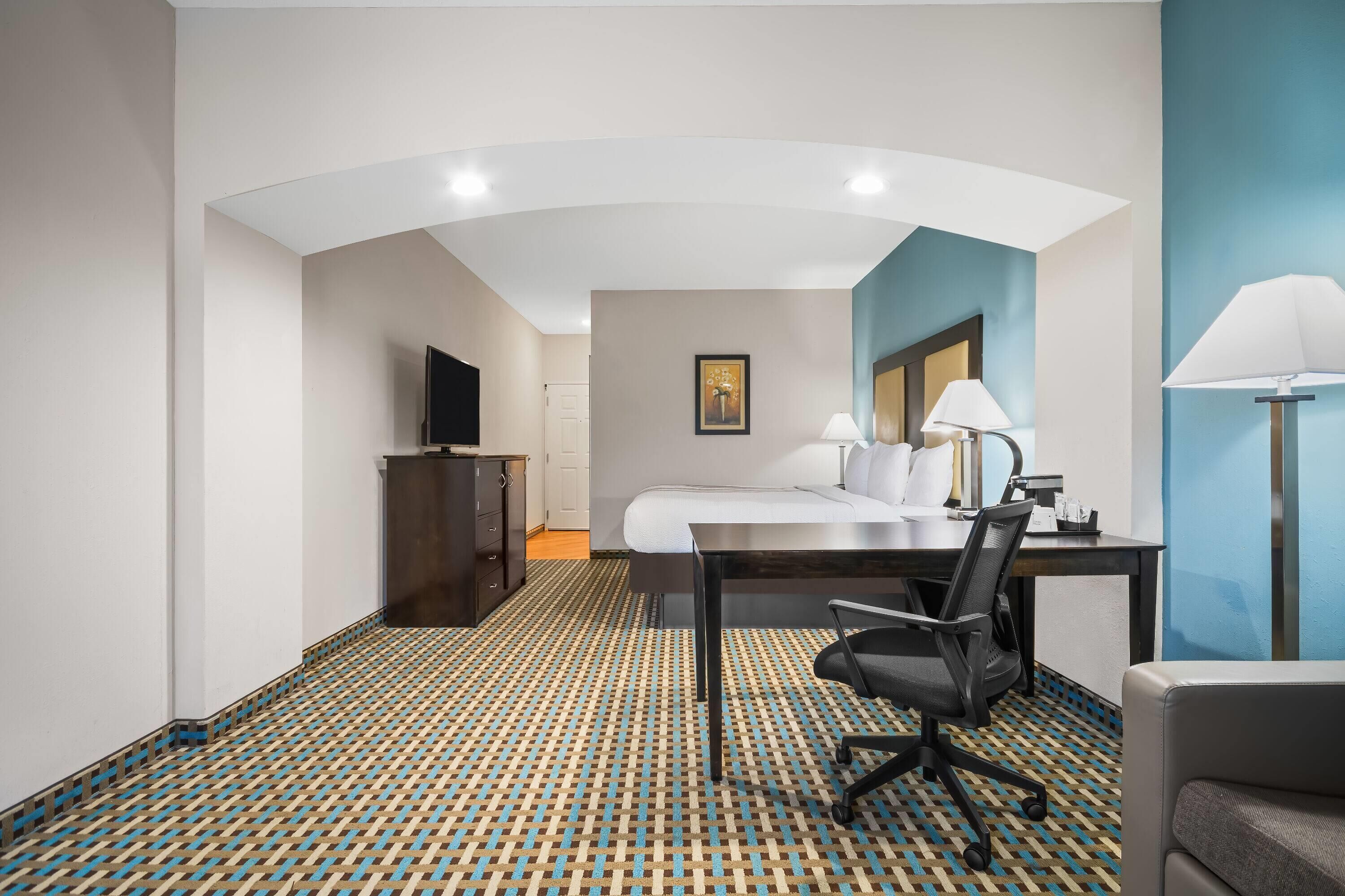 Habitación de hotel con cama, escritorio, silla, lámpara, televisión y alfombra estampada.