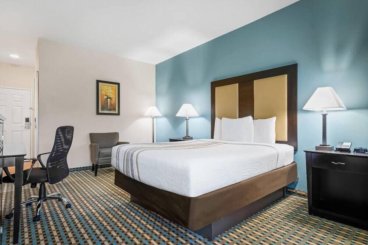 A hotel room with a bed, desk, two chairs, a desk lamp, a bedside table, and a wall-mounted mirror.