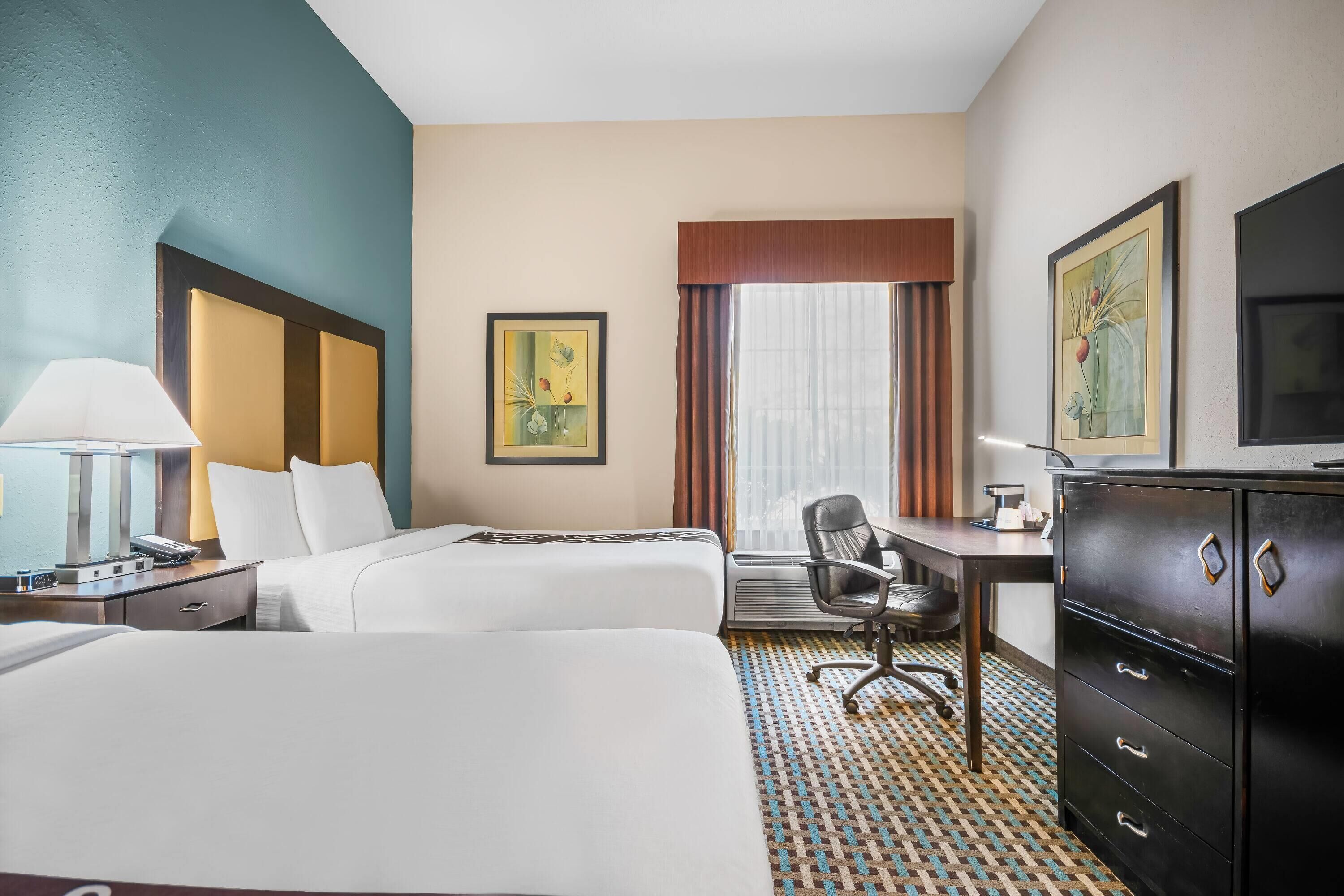 A hotel room with two beds, a desk, a chair, a dresser, and artwork on the walls.