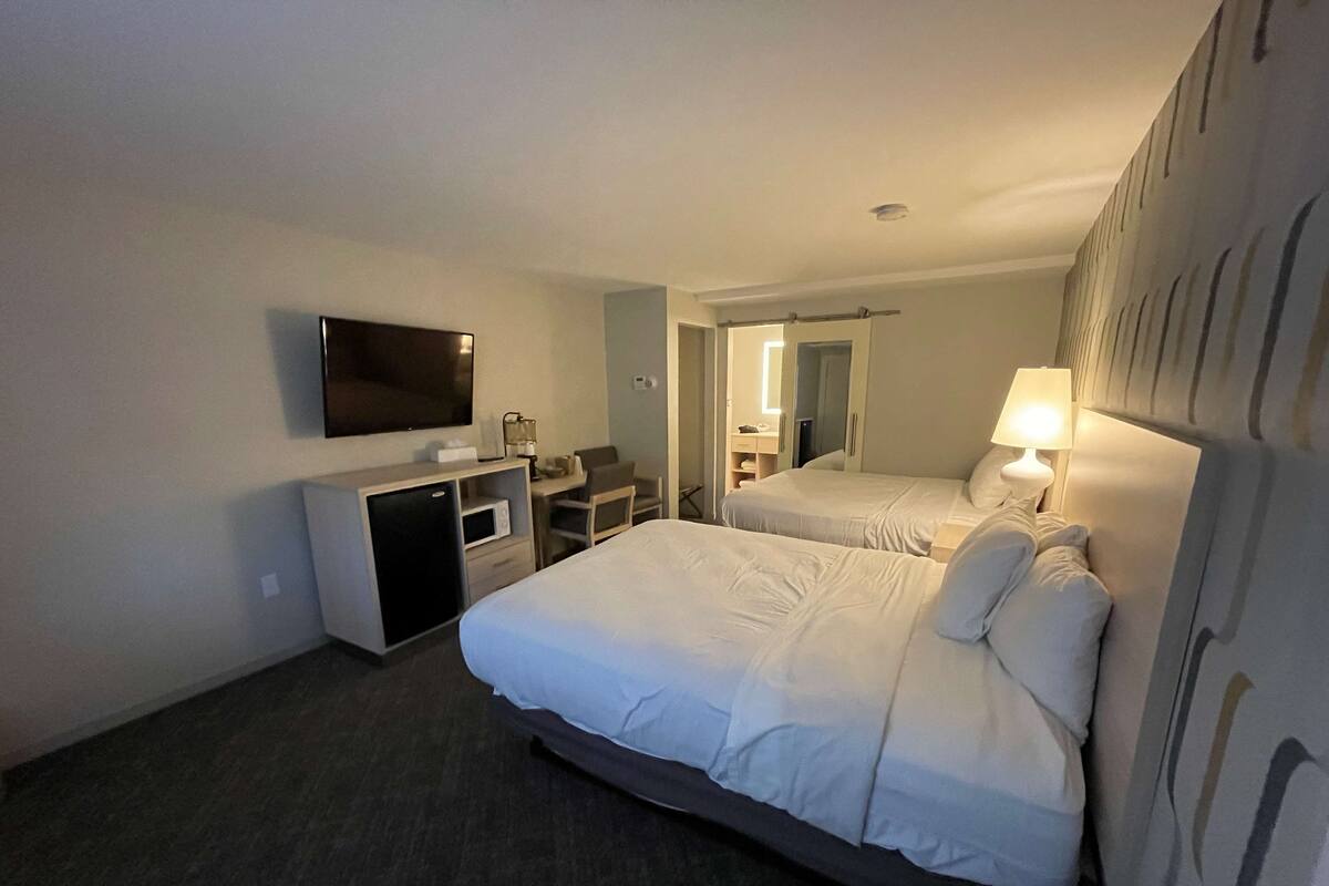 A hotel room with two beds, a desk, a TV, and a small kitchenette.