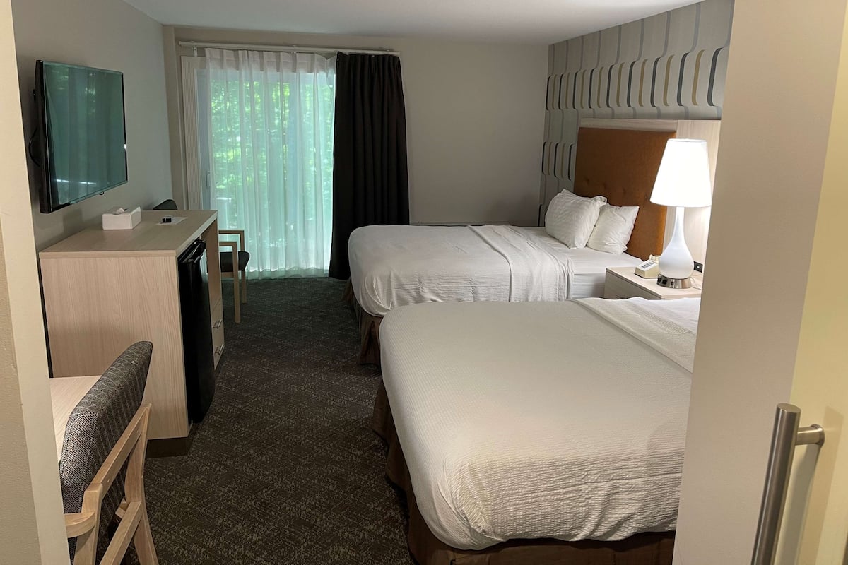 A hotel room with two beds, a desk with a TV, a chair, a window with curtains, and a lamp.