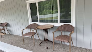 A patio with two wooden tables and four wicker chairs, set against a grey wooden wall with a large window above.