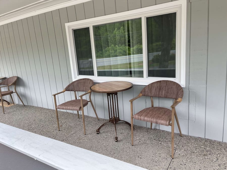 A patio with two wooden tables and four wicker chairs, set against a grey wooden wall with a large window above.