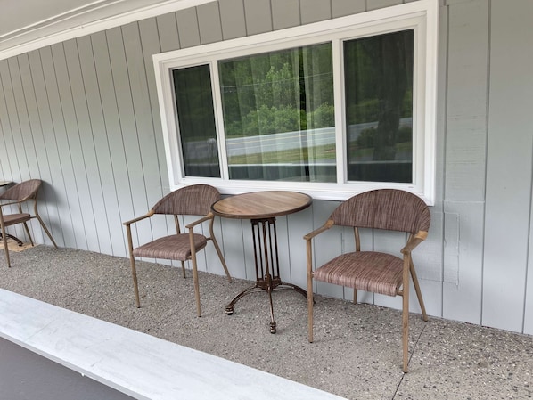 Une terrasse avec deux tables en bois et quatre chaises en osier, adossée à un mur en bois gris surmonté d’une grande fenêtre.