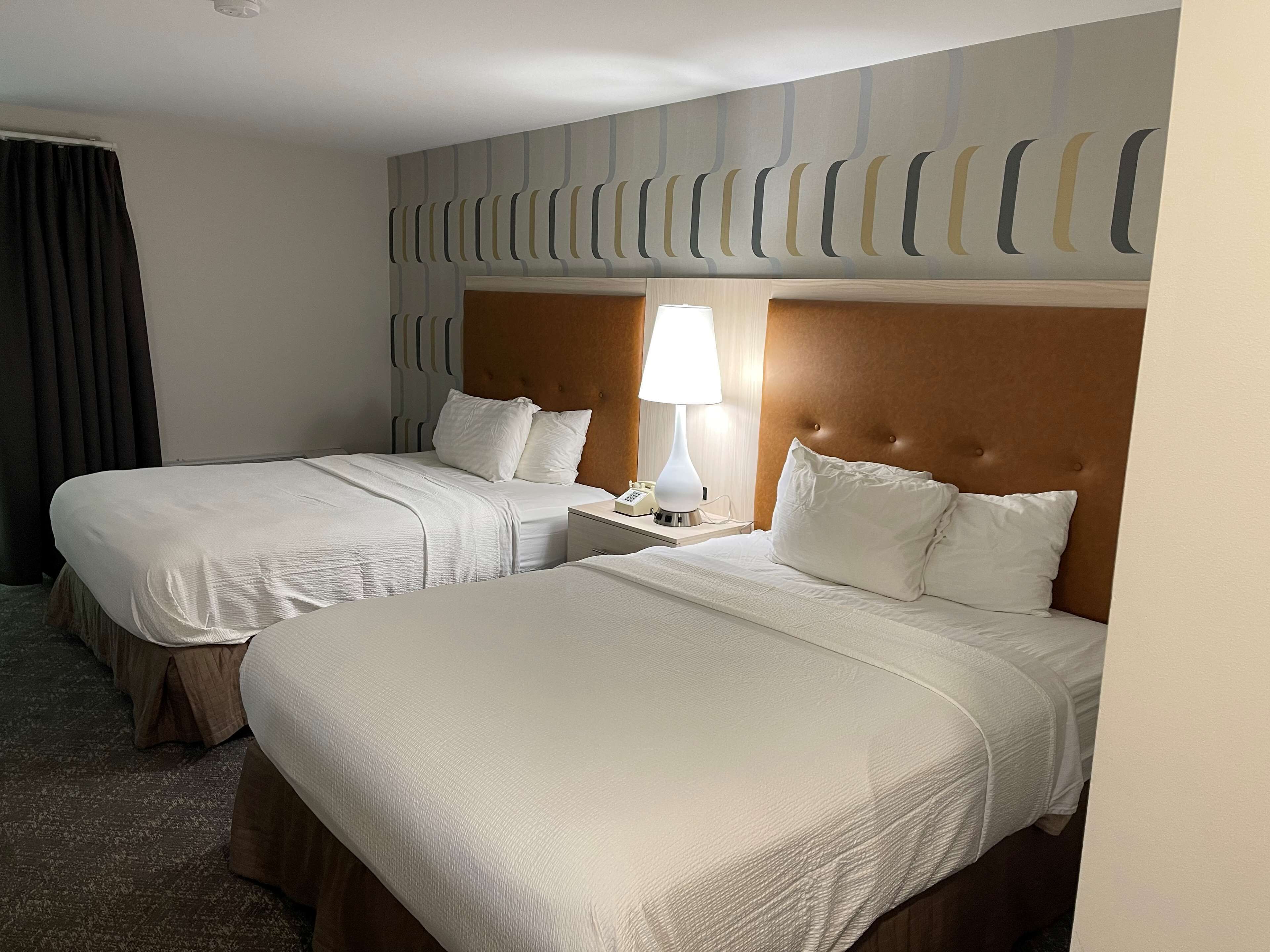 A hotel room with two beds, each with white linens and a wooden headboard. There is a bedside table with a lamp and a phone.
