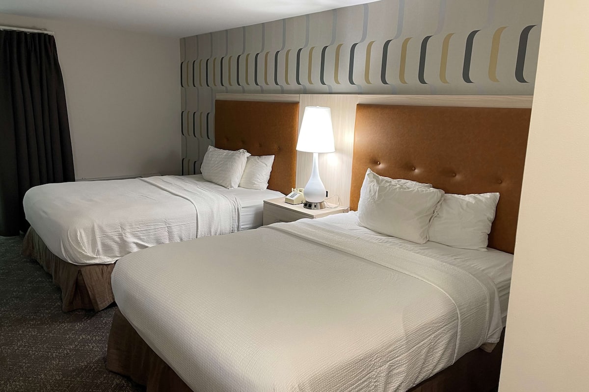 A hotel room with two beds, each with white linens and a wooden headboard. There is a bedside table with a lamp and a phone.