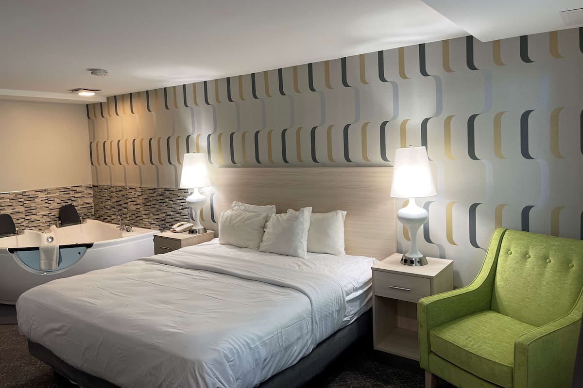 A hotel room with a bed, two bedside tables, a green armchair, a bathroom with a bathtub, and a patterned wall.