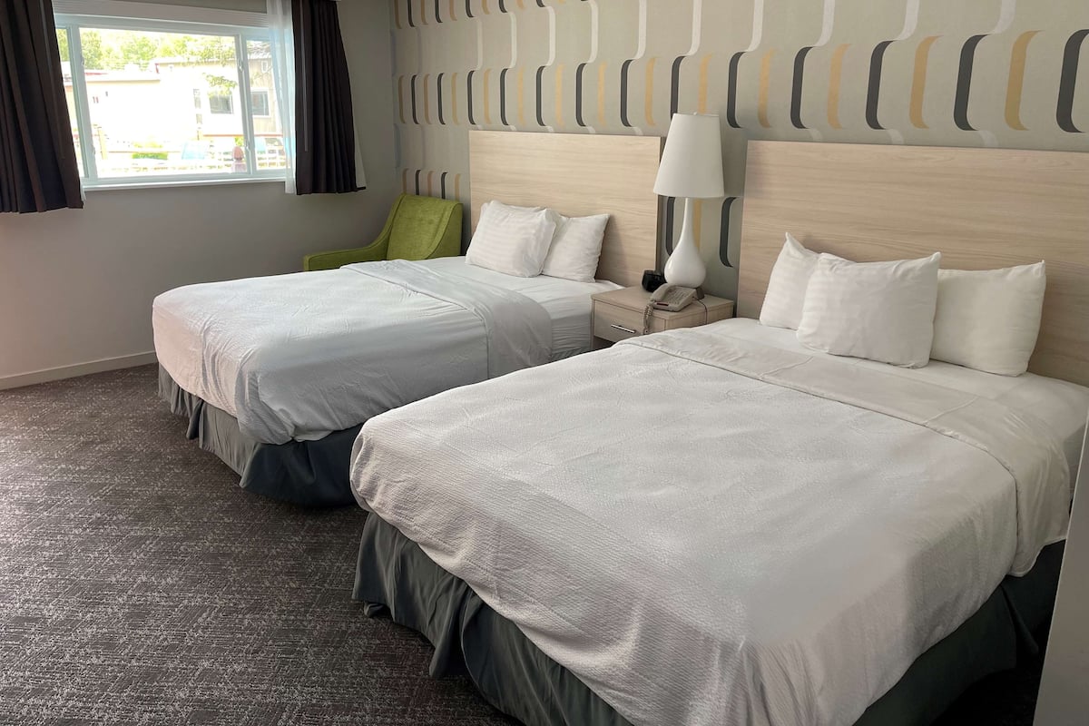 A hotel room with two beds, a window with curtains, a green chair, a bedside table with a lamp, and a patterned wall.