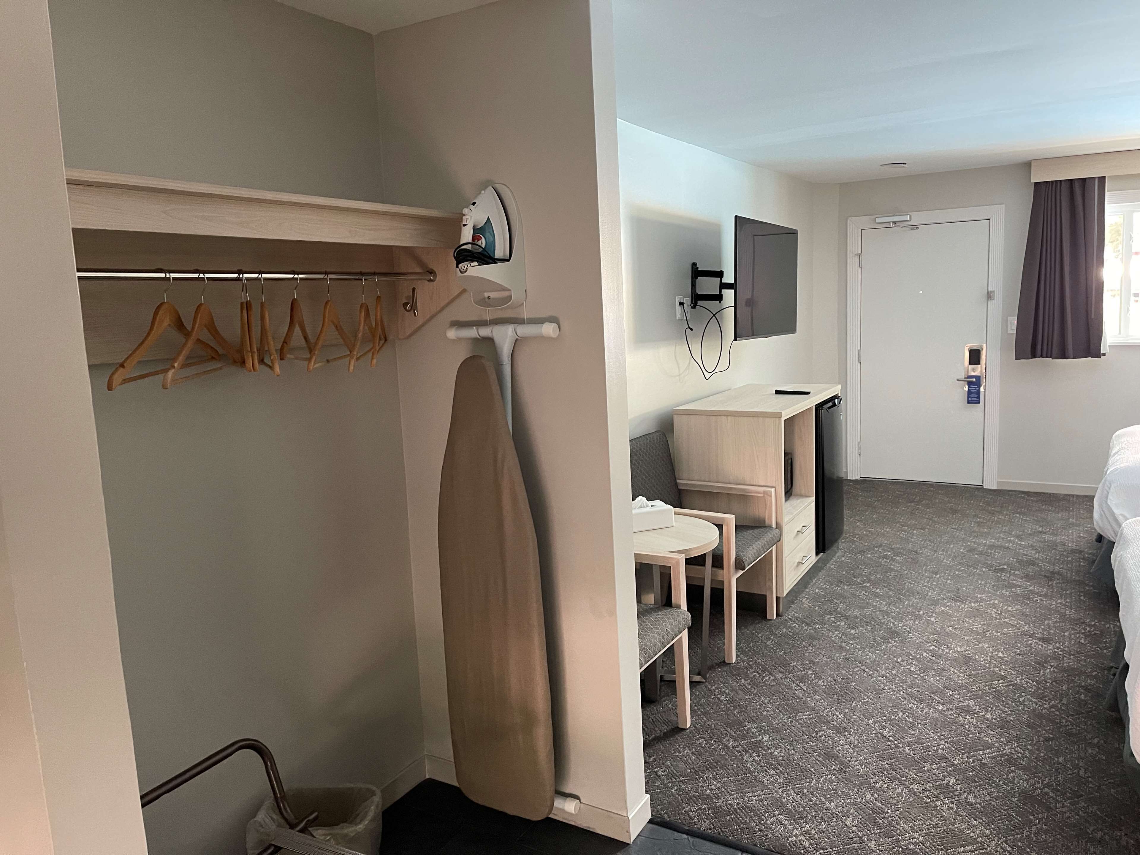 A hotel room with a bed, a desk with a chair, a TV, and a coat rack with hangers.